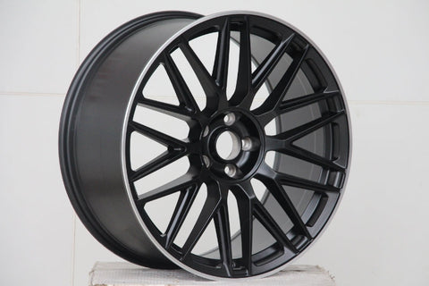 A matte black alloy wheel with a multi-spoke design is resting on a white, textured surface, set against a plain white background.