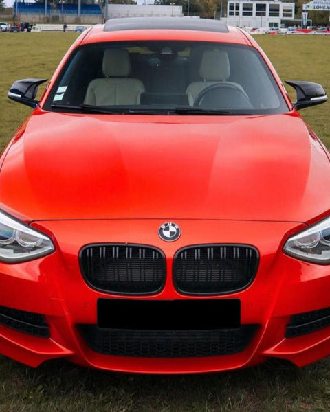 A bright orange car, stationary, is parked on a grassy field. It features a distinctive grille with a BMW emblem and sleek headlights, surrounded by an open area with distant buildings.