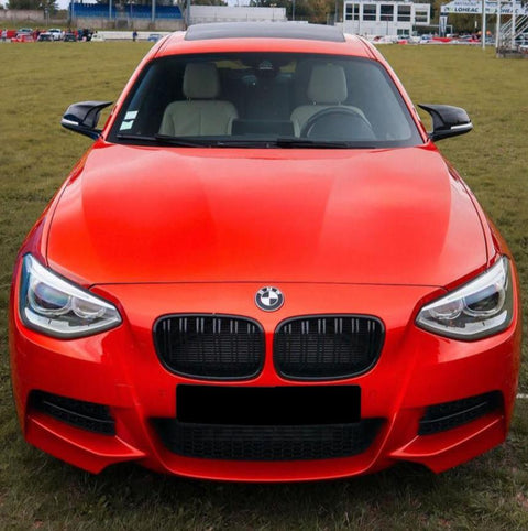 A bright orange car, stationary, is parked on a grassy field. It features a distinctive grille with a BMW emblem and sleek headlights, surrounded by an open area with distant buildings.