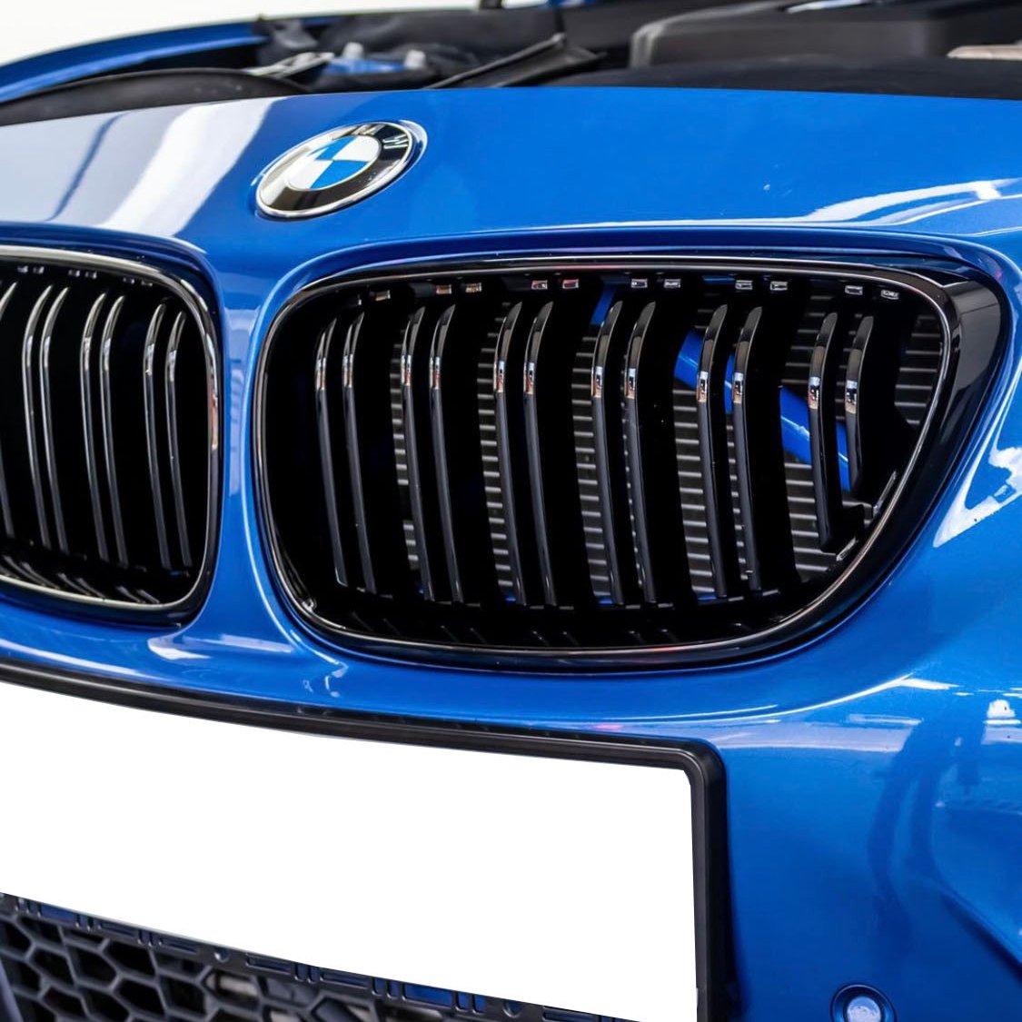 A blue car features a black kidney grille and BMW logo, with an open hood visible in a garage setting. The license plate area is blank.