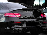 A black Mercedes-Benz AMG C63 parked, with illuminated rear lights, chrome quad exhausts, and badges. It is set against a modern building with reflective windows and dim lighting.