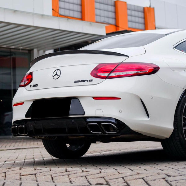 A white Mercedes-Benz C 300 AMG is parked on a cobblestone surface. The car's rear features dual exhausts and red tail lights, with an orange and white building in the background.