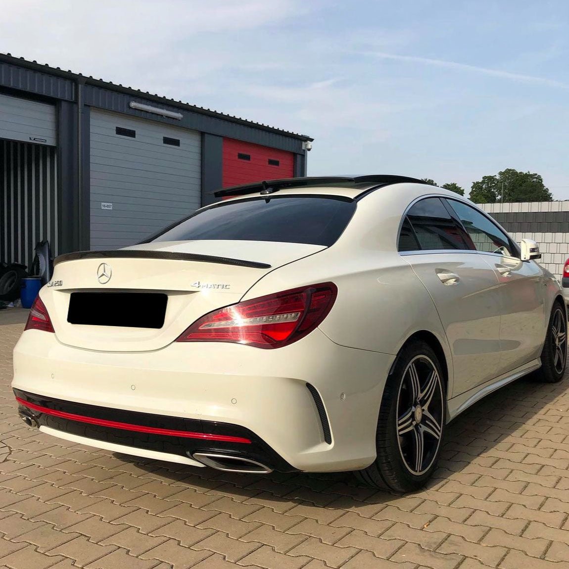 A white Mercedes-Benz CLA 250 4MATIC car is parked on a cobblestone driveway. Background includes a garage with red and gray doors.