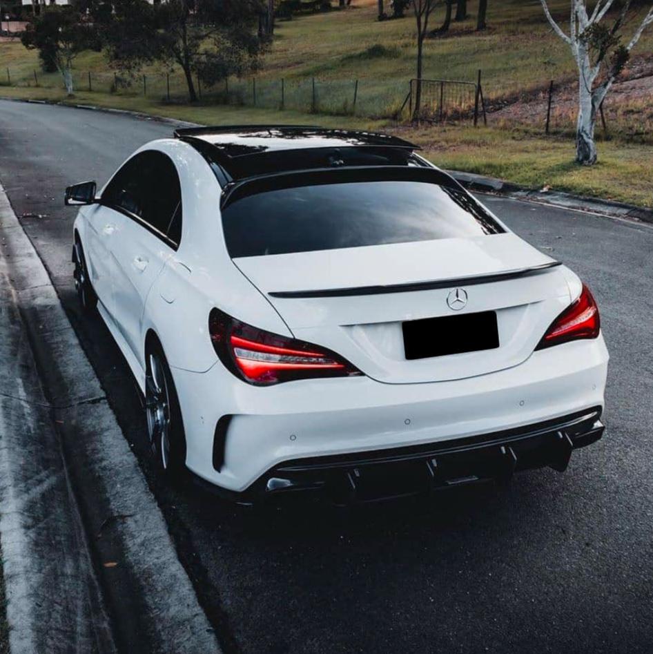 A white car with sleek design and illuminated taillights is parked on a street next to a grassy area with trees in the background.
