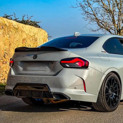 A silver car with a spoiler is parked on a road, near a stone wall and a leafless tree, under a blue sky.