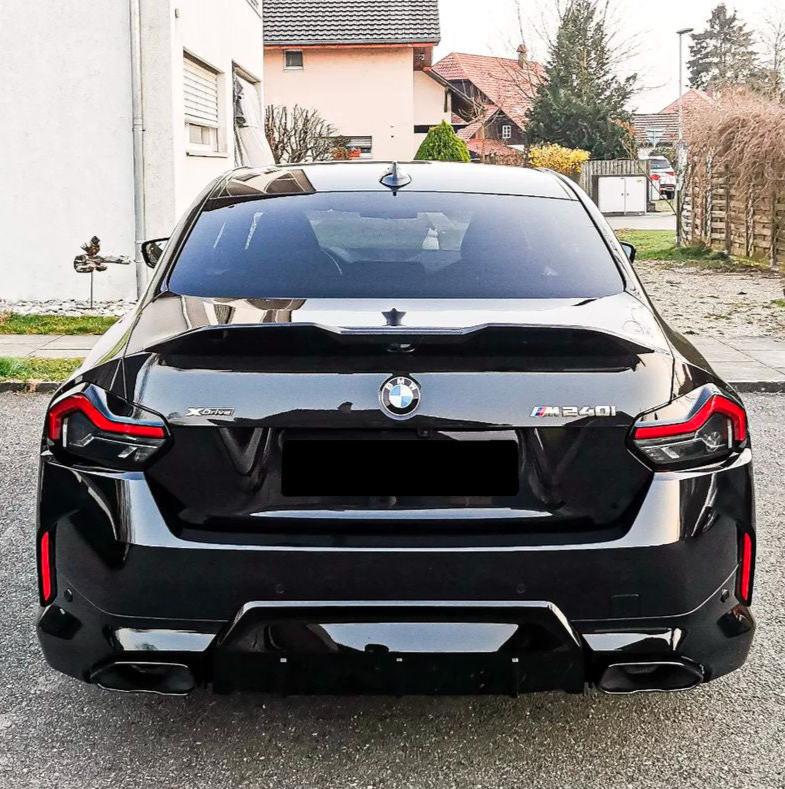 A black BMW M240i is parked on a driveway, surrounded by residential houses and green lawn. The car's prominent features include taillights and visible badges reading "X Drive" and "M240i".