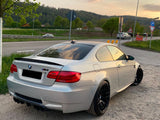 A silver BMW M3 is parked on a gravel area beside a road, with a backdrop of greenery and trees under a partly cloudy sunset sky.