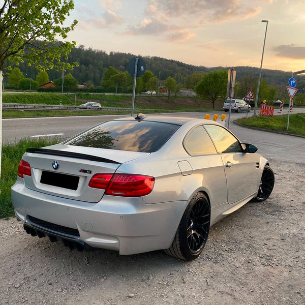 A silver BMW M3 is parked on a gravel area beside a road, with a backdrop of greenery and trees under a partly cloudy sunset sky.