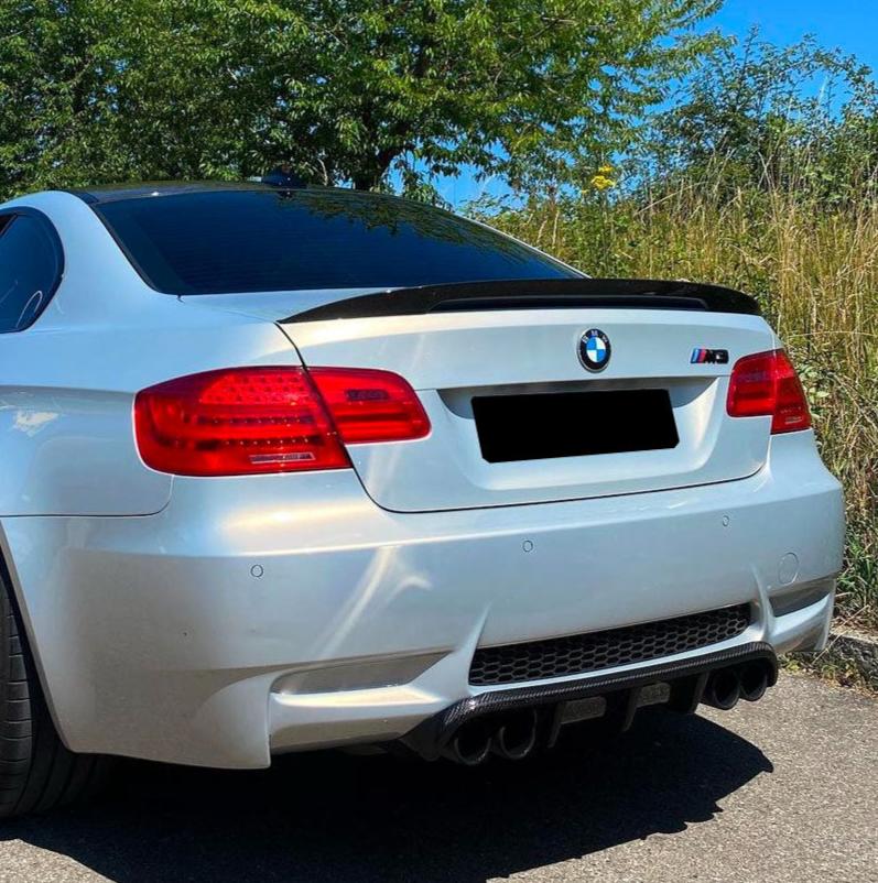A white BMW M3 is parked on the street, surrounded by tall grass and trees. The car's rear view showcases its taillights and four exhaust pipes.