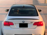 A white BMW M3 is parked on a sunlit pavement. The rear view shows its dual exhausts, taillights, and the BMW emblem. A modern building with poles is visible in the background.