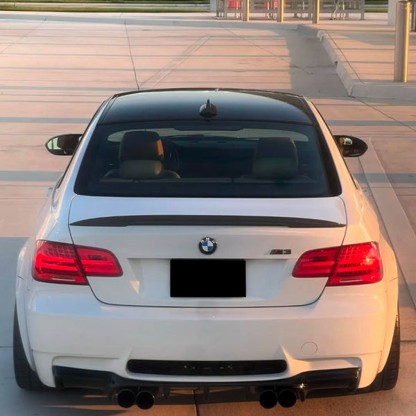A white BMW M3 is parked on a sunlit pavement. The rear view shows its dual exhausts, taillights, and the BMW emblem. A modern building with poles is visible in the background.