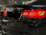 A black BMW M3 with illuminated red taillights is parked indoors. The dual exhaust and carbon fiber bumper highlight its sporty design. A blurred workshop setting surrounds the car.