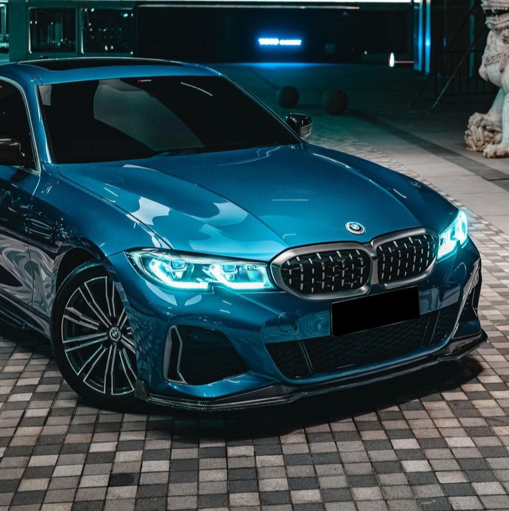 A blue car with illuminated headlights is parked on a checkered pavement beside a building and a statue, under ambient lighting.