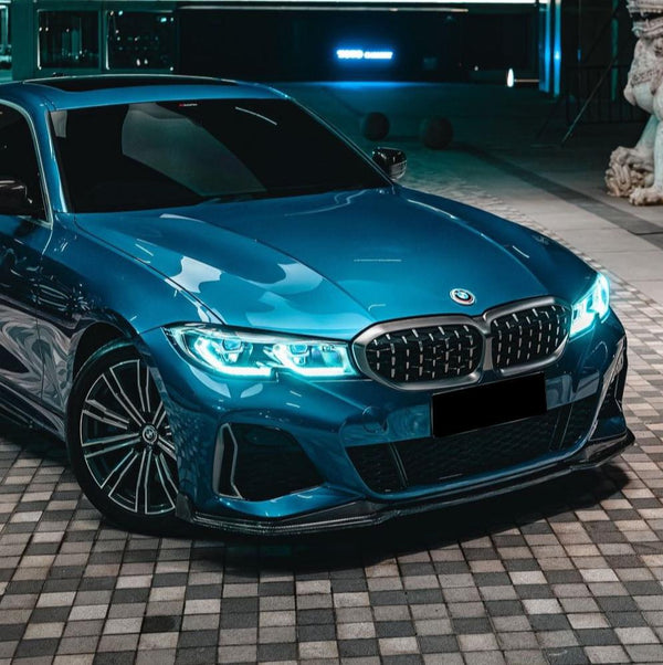 A blue car with illuminated headlights is parked on a checkered pavement beside a building and a statue, under ambient lighting.