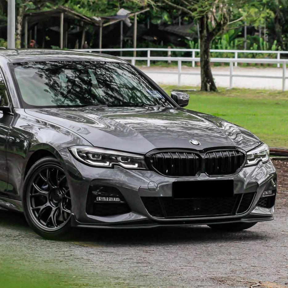 A sleek gray car, parked on a paved area, features distinctive front grille and sharp headlights, surrounded by lush green grass and trees.