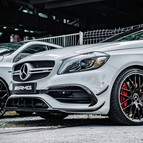 A white Mercedes-Benz AMG car is parked on a concrete surface, featuring a prominent grille and sleek wheel design. A fence and an industrial structure are in the background.