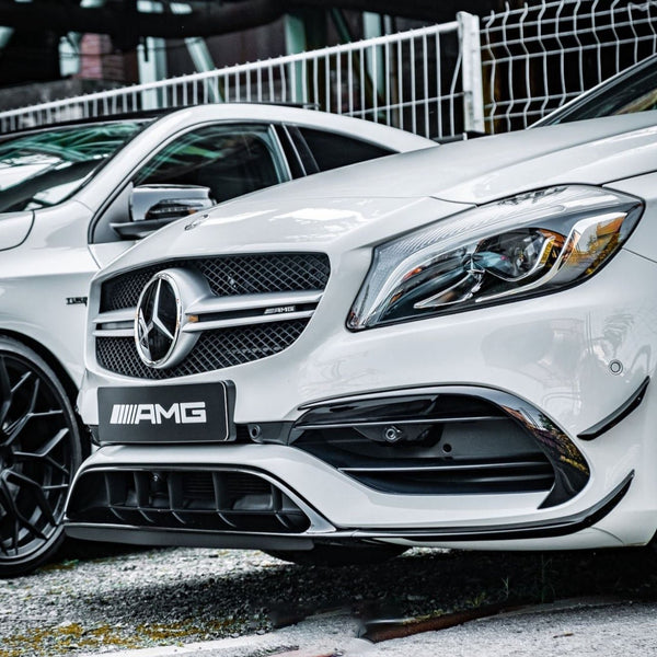 A white Mercedes-Benz car, featuring the AMG badge, is parked on a gravel surface near a mesh fence, highlighted by the sleek front design and prominent logo.