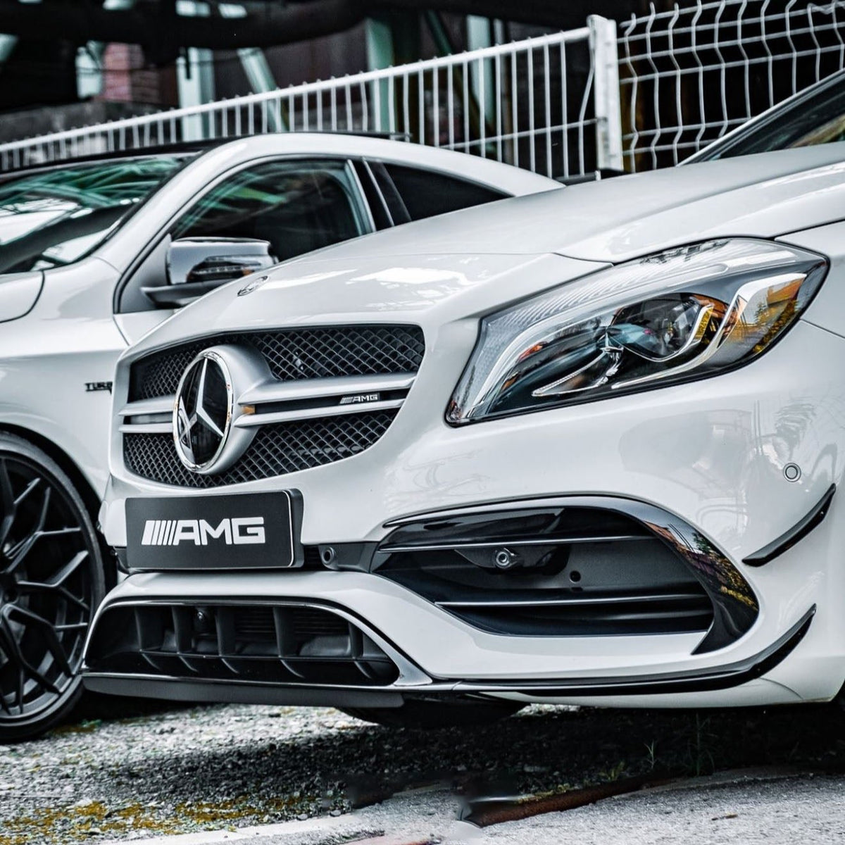 A white Mercedes-Benz car, featuring the AMG badge, is parked on a gravel surface near a mesh fence, highlighted by the sleek front design and prominent logo.