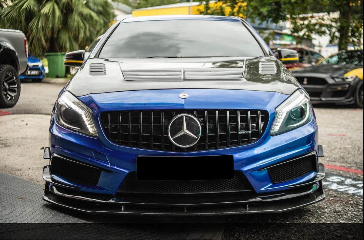 A blue Mercedes-Benz car is parked on a street, featuring a blacked-out grille and hood with vents. Surrounding it are trees and other parked vehicles in an outdoor setting.