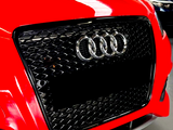 Red car grille features the silver Audi logo with four rings, surrounded by a honeycomb pattern, set in an indoor environment alongside other parked cars.