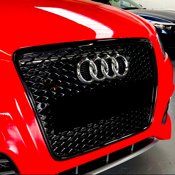 Red car grille features the silver Audi logo with four rings, surrounded by a honeycomb pattern, set in an indoor environment alongside other parked cars.