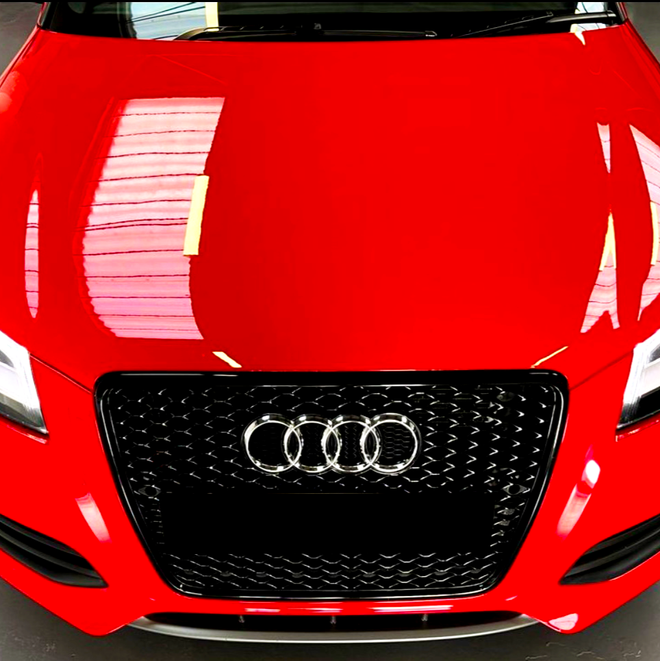 A red car's front view, displaying a black grille with four interlocking silver rings, is parked indoors with light reflections visible on its hood.