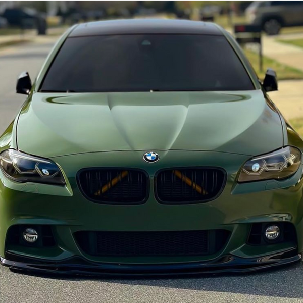 A matte green BMW is parked on a suburban street, featuring tinted windows and a lowered stance, with houses and a driveway visible in the background.