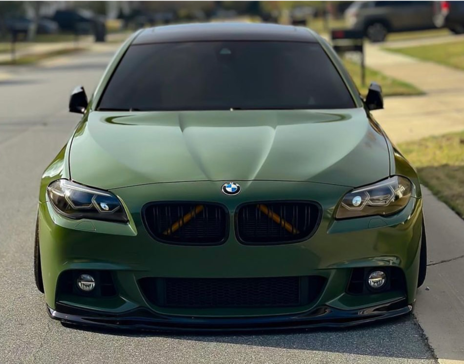 A matte green BMW is parked on a suburban street, featuring tinted windows and a lowered stance, with houses and a driveway visible in the background.
