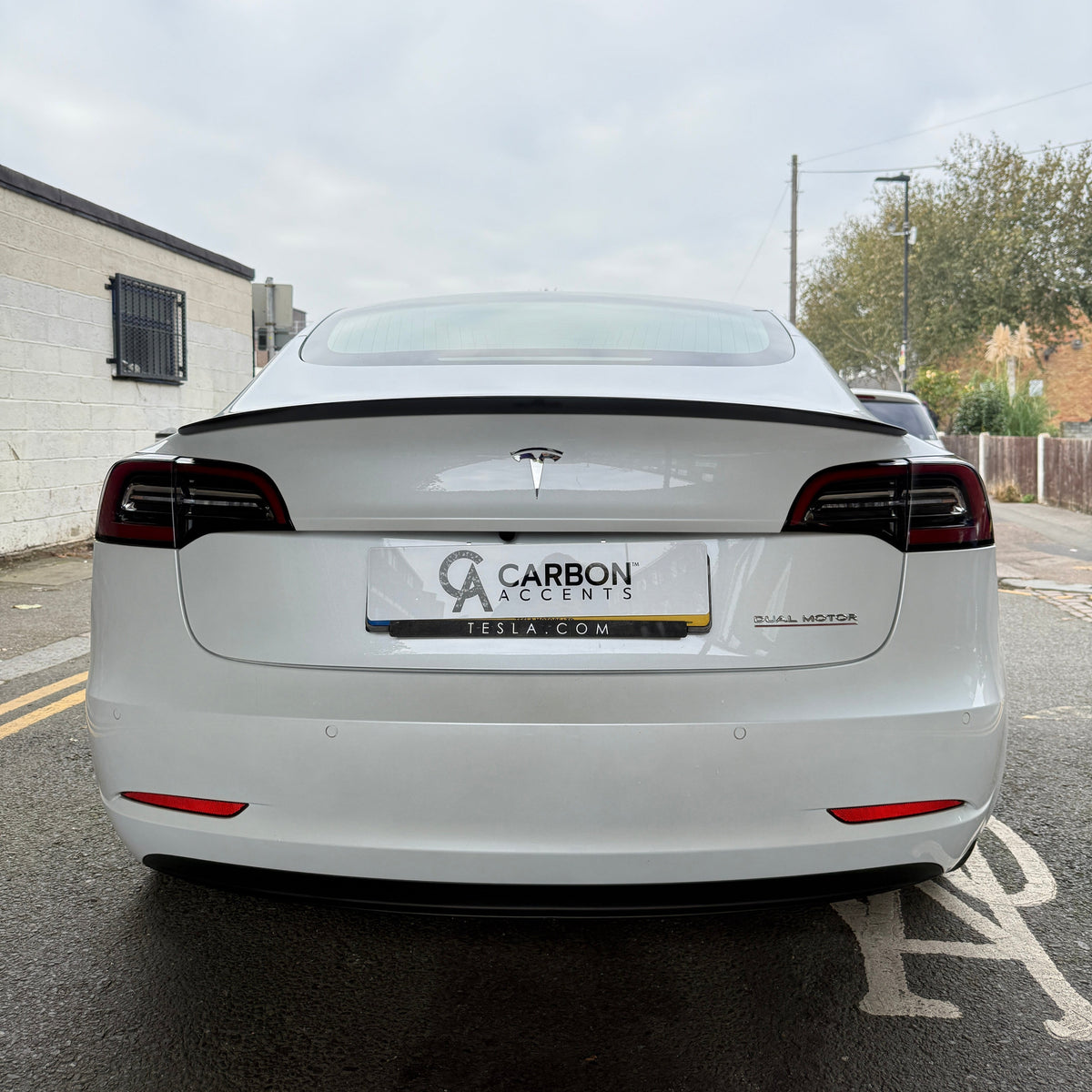 A white Tesla car is parked, displaying "CARBON ACCENTS" and "TESLA.COM" on its license plate. The words "DUAL MOTOR" appear on the rear. It's positioned on a street near buildings.
