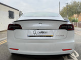 A white Tesla Model with dark-tinted tail lights is parked in an urban street. The license plate reads 