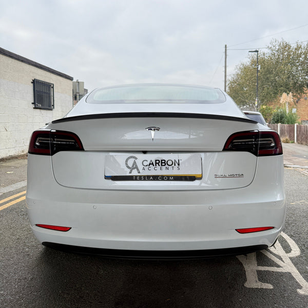 A white Tesla Model with dark-tinted tail lights is parked in an urban street. The license plate reads "CARBON ACCENTS TESLA.COM." There's a building and trees in the background.