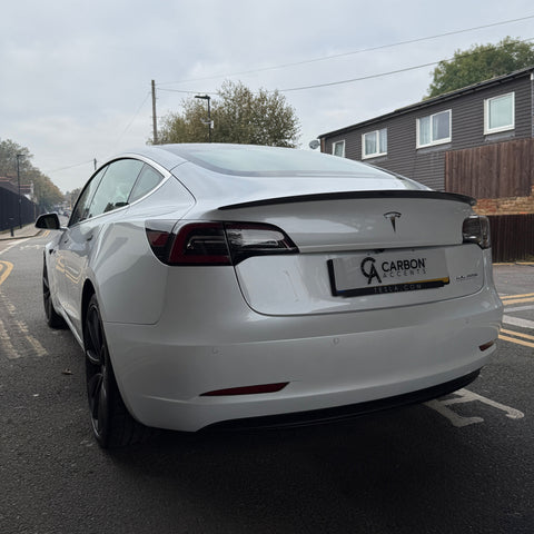 A white Tesla car is parked on a street near residential buildings. Text reads "CARBON ACCENTS," and "TESLA.COM" on the license plate frame. Overcast sky overhead.