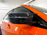 A sleek, black side mirror is attached to an orange car, reflecting light in a well-lit indoor garage setting.
