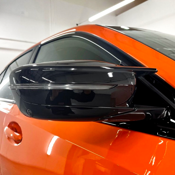 A sleek, black side mirror is attached to an orange car, reflecting light in a well-lit indoor garage setting.