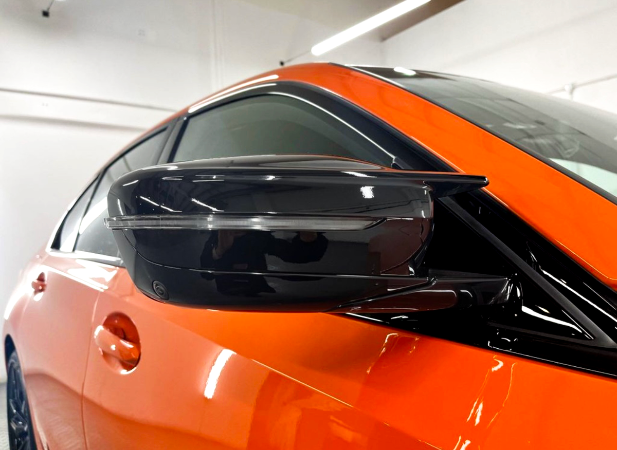A sleek, black side mirror is attached to an orange car, reflecting light in a well-lit indoor garage setting.