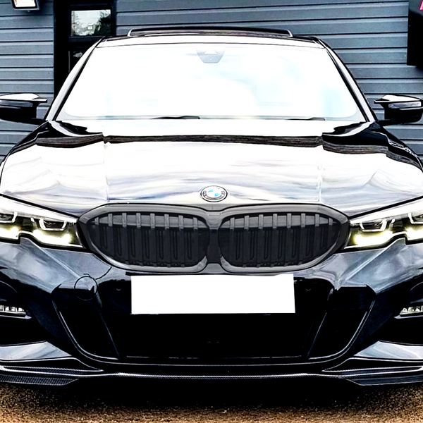 A black BMW car is parked with its headlights on, facing forward. In the background, there's a gray building with a sign reading "A6 MOTORS SPECIALIST VEHICLE PROVIDER."