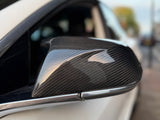 Car side mirror with a carbon fiber cover, reflecting light, attached to a white vehicle. The background includes a blurred street scene with a pedestrian walkway.