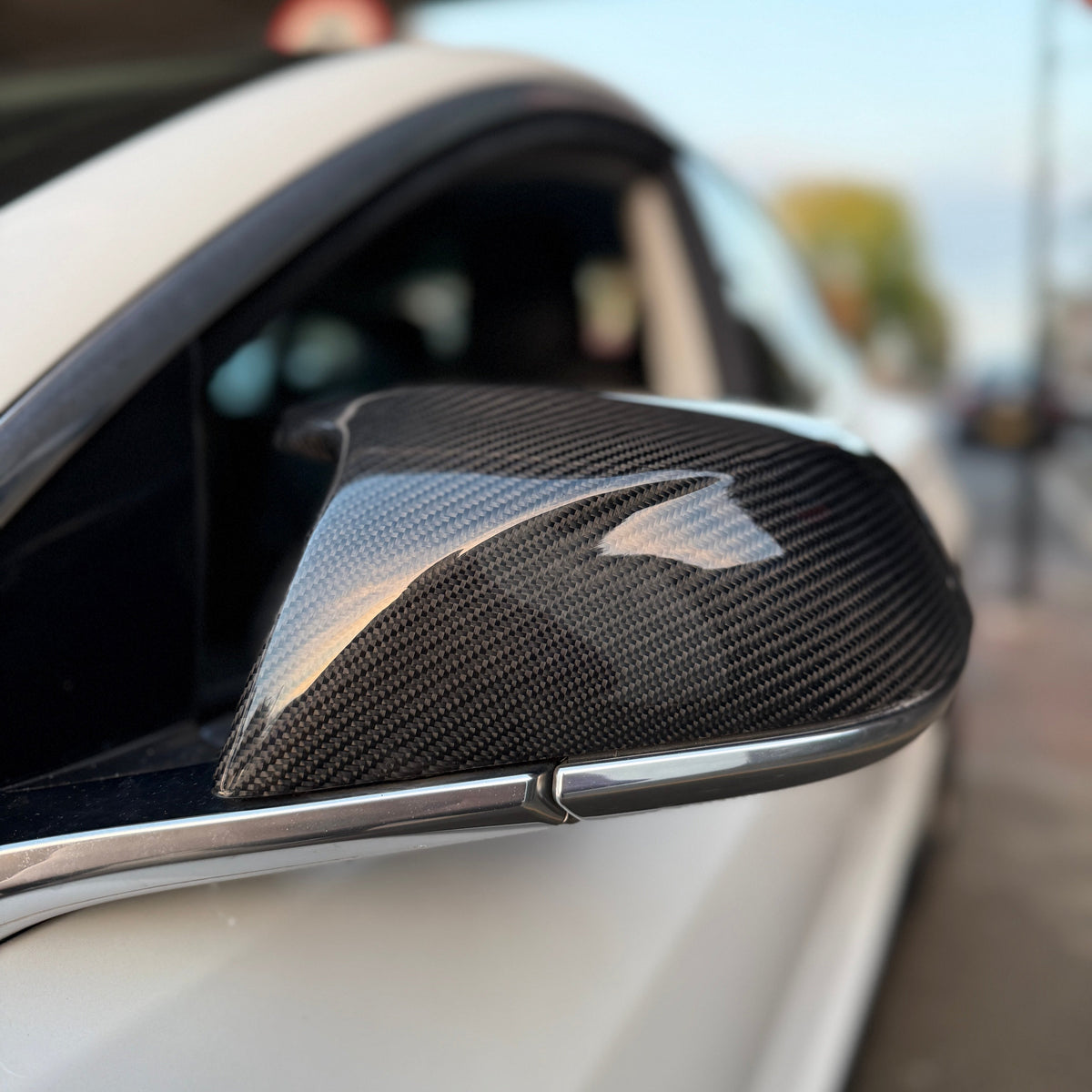 Car side mirror with a carbon fiber cover, reflecting light, attached to a white vehicle. The background includes a blurred street scene with a pedestrian walkway.