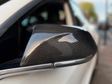 A carbon fiber wing mirror is attached to a white car, reflecting light in an urban street setting with blurred background.