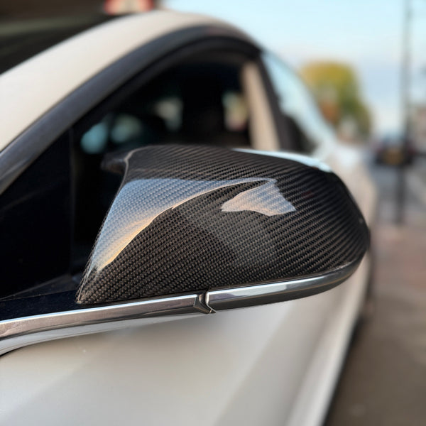 A carbon fiber wing mirror is attached to a white car, reflecting light in an urban street setting with blurred background.