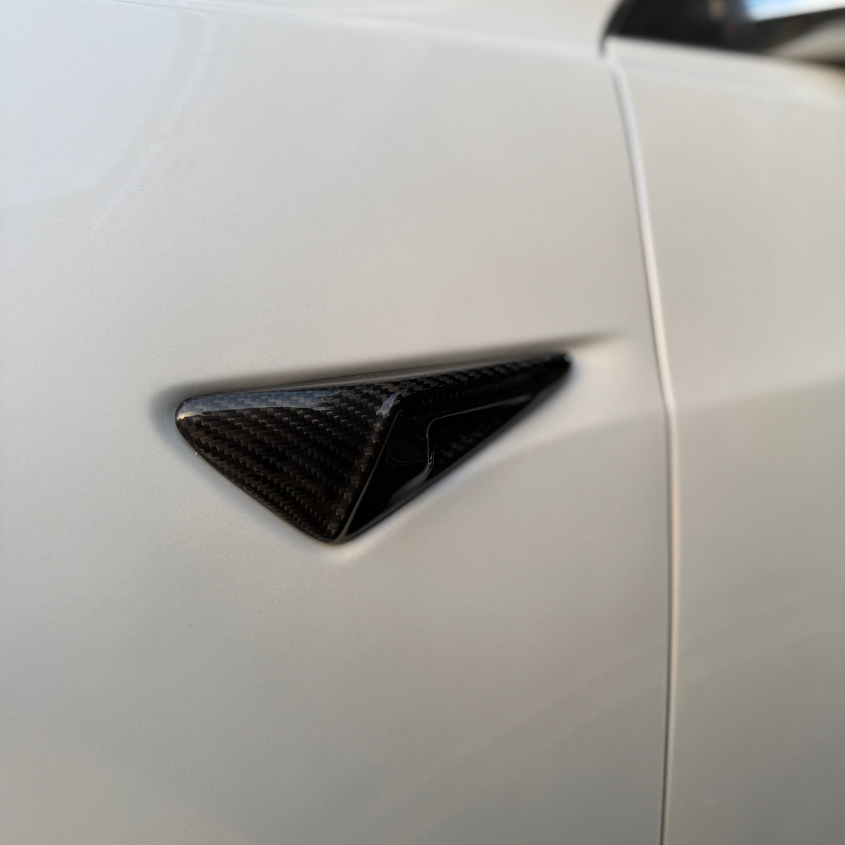 A carbon fiber car door handle resides on a smooth, light-colored vehicle door, surrounded by softly blurred ambient light, enhancing its sleek and modern design.
