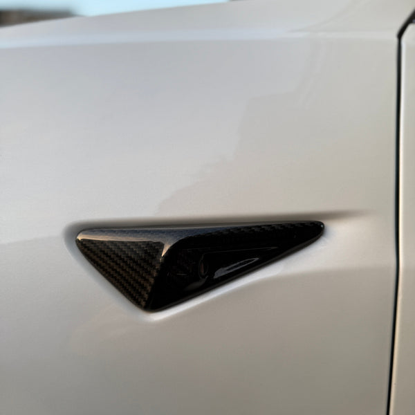 A carbon fiber side camera, embedded in a sleek, white car's body, is situated near the edge, beneath the side mirror. The setting is outdoors with soft lighting.