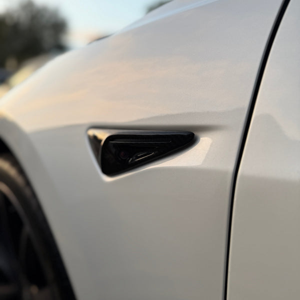 A sleek front fender vent with a glossy black finish is mounted on a shiny white car, reflecting soft ambient light, set outdoors.