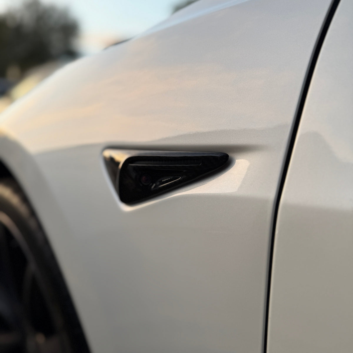 A sleek, white vehicle door with a black, triangular side marker integrates into the curved body. The background is blurred, suggesting an outdoor setting with soft lighting.