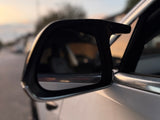 A side-view mirror reflects blurred surroundings on a parked vehicle during sunset, with a soft-focus background of a quiet street and distant trees.