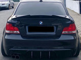 A black BMW car is parked on a residential street. The rear is prominently featured, displaying tinted windows and dual exhaust pipes. Nearby is another parked vehicle and houses.