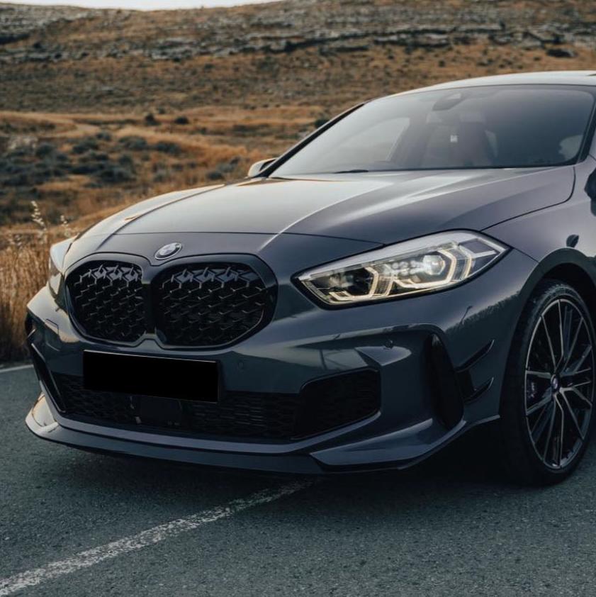 A sleek gray car with illuminated headlights is parked on a road, surrounded by a rugged, grassy landscape. The front grille is distinctive, and the wheels feature intricate designs.