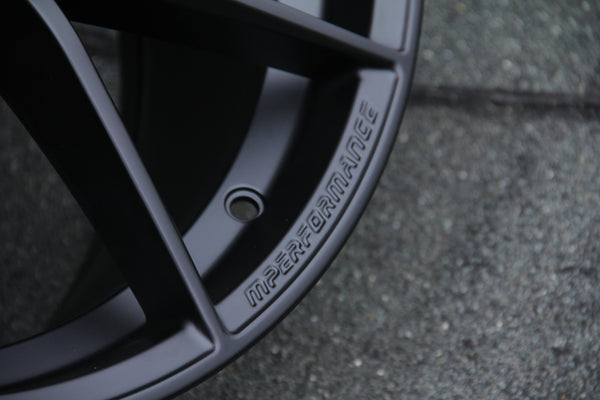 A black wheel rim is shown with embossed text reading "MPERFORMANCE," set against a textured gray surface in a close-up, highlighting its sleek design.