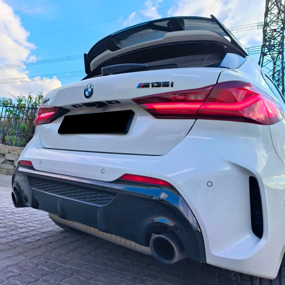 A white BMW M135i with an open rear hatch, parked on a cobblestone surface, surrounded by greenery and electrical towers under a blue sky.