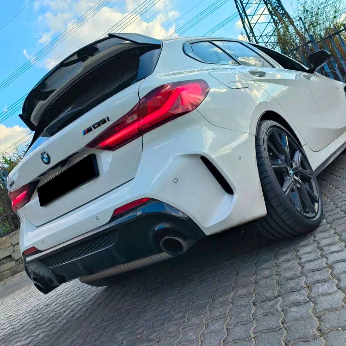 A white BMW M135i with a slightly open trunk is parked on a cobblestone surface, surrounded by a fence and power lines under a blue sky.